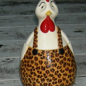 Puede incluir: Una figurita de gallo de cer&aacute;mica con cuerpo blanco, cresta y barbilla rojos, y un estampado de leopardo marr&oacute;n y negro en su mono.