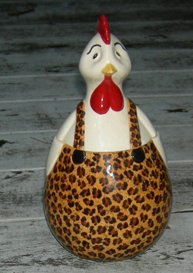 Puede incluir: Una figurita de gallo de cer&aacute;mica con cuerpo blanco, cresta y barbilla rojos, y un estampado de leopardo marr&oacute;n y negro en su mono.