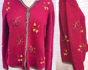May include: A burgundy cardigan sweater with a V-neck and button closure. The sweater features embroidered leaf and berry designs in yellow, brown, and gold. The trim and button details are brown.