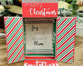 May include: A red and green striped paper box with a red top that says "merry Christmas". The box is open and inside is a white card with the quote "Joy comes from, & because of Him." by President Russell M. Nelson. The bottom of the box has a white silhouette of the nativity scene.