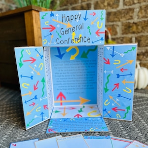 May include: A blue and white cardboard box with a pop-up design. The box has a blue background with colorful arrows and the words "Happy General Conference" on the front. The inside of the box has a quote from the Book of Doctrine and Covenants.