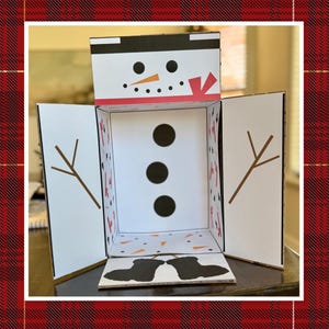 Könnte beinhalten: Eine weiße Papp-Schneemann-Box mit einem schwarzen Zylinder, einer orangefarbenen Nase und schwarzen Augen. Die Box ist geöffnet und zeigt ein weißes Inneres mit drei schwarzen Knöpfen und einem braunen Zweig auf jeder Seite. Der Boden der Box hat schwarze Stiefel.