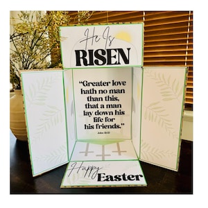以下が含まれることがあります： グリーンとホワイトの紙製のイースターカードで、ポップアップデザインになっています。カードには「He is RISEN」という文字と、ヨハネ13:13からの引用「Greater love hath no man than this, that a man lay down his life for his friends.」が書かれています。カードには緑色の十字架と「Happy Easter」の文字も含まれています。