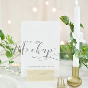 May include: A white table sign mockup with the text "Table Sign Mockup 5x7" on a wooden base. A lit white candle and greenery are in the background. The text "Physical items are not for sale" is at the bottom.