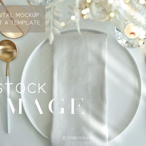 May include: A white dinner plate with a gold rim, a white linen napkin, a gold fork and spoon, and a clear glass candle holder with a white candle. The plate is on a white surface.