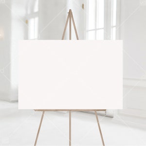 May include: A white canvas on a wooden easel, standing in a white room with a window in the background.