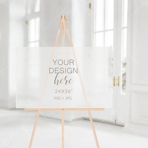 以下が含まれることがあります： 木製のイーゼルに置かれた白いアクリル製の看板。看板には「YOUR DESIGN here 24x36」PSD + JPGと書いてあります。