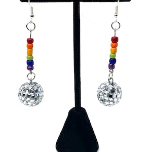 May include: Dangle earrings featuring a rainbow bead design and a disco ball charm. The earrings have a silver hook and a series of colourful beads in red, orange, yellow, green, blue, and purple. The disco ball charm is made of small, reflective squares.