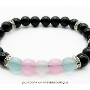 Könnte beinhalten: Ein Perlenarmband mit glatten, schwarzen, kugelförmigen Perlen, unterbrochen von silberfarbenen Zwischenelementen mit kleinen, klaren Kristallen. In der Mitte befinden sich zwei hellblaue, zwei rosa und eine weiße, durchscheinende Perle. Text: @UNICORNWHISPERERBOUTIQUE.