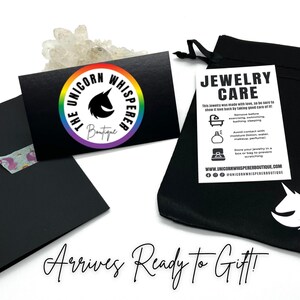 May include: A black drawstring jewellery pouch with a white unicorn logo, a black business card featuring a rainbow circular logo with the text The Unicorn Whisperer Boutique, and a white jewellery care instruction card with black text.