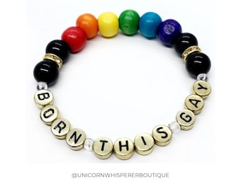 Può includere: Un braccialetto di perline con una sequenza di perline color arcobaleno, perline nere e perline distanziatrici dorate. Il braccialetto include perline con lettere dorate che compongono la frase BORN THIS GAY. Il testo @UNICORNWHISPERERBOUTIQUE è visibile.
