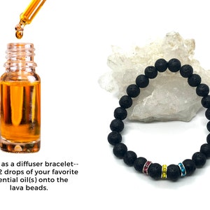 A black lava rock bracelet with pink, yellow and blue rhinestones. The bracelet is on a white surface. A bottle of essential oil and a dropper are shown to the left of the bracelet. Text reads: 'To use as a diffuser bracelet-- add 1-2 drops of your favorite essential oil(s) onto the lava beads.'