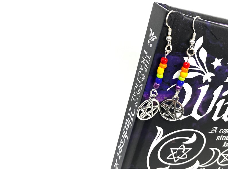 May include: A pair of drop earrings featuring silver-toned pentacle charms suspended from silver hooks. Each earring includes a vertical row of small, vibrant glass beads in a rainbow sequence of red, orange, yellow, green, blue, and purple.