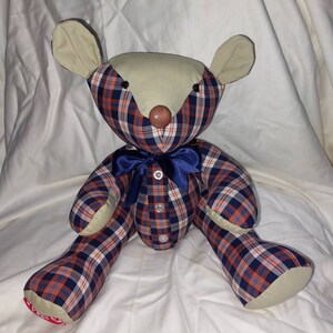 May include: A handmade plaid teddy bear with a blue bow tie. The bear is made from a blue, red, and white plaid fabric. The bear has a beige face and brown button eyes.