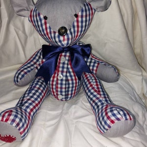 May include: A gray and white plaid teddy bear with a blue bow tie. The bear is sitting on a white surface.