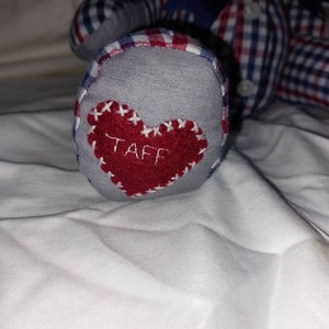 May include: A gray fabric foot with a red heart sewn on it. The heart has the word "TAFF" embroidered in white thread.