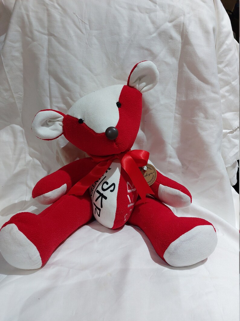 May include: Red and white teddy bear with a red ribbon around its neck. The bear has a brown nose and black eyes. The bear is sitting on a white surface. The bear has the words "SKP" and "Love you always" written on its chest.