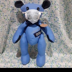 May include: Blue teddy bear wearing a white face mask. The bear has black ears and a University of Hertfordshire logo on its chest.
