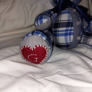 May include: A blue and white plaid teddy bear with a red heart on its foot. The heart has a white letter G stitched on it.