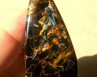 May include: A teardrop-shaped cabochon of blue, gold, and brown tiger's eye gemstone. The stone has a unique pattern of swirling colors and a high polish.