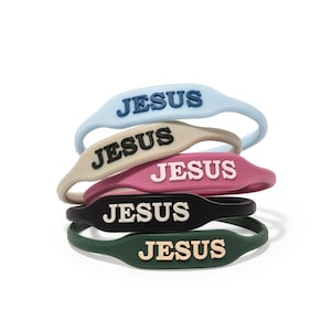 May include: A stack of five silicone wristbands in various colors: light blue, tan, pink, black, and dark green. Each band features the word "JESUS" in a contrasting color. The bands are stacked on a white background.