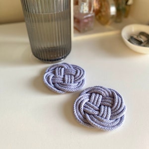 May include: Two lavender-coloured, round, woven rope coasters. The coasters are made of thick rope and have a knot design.