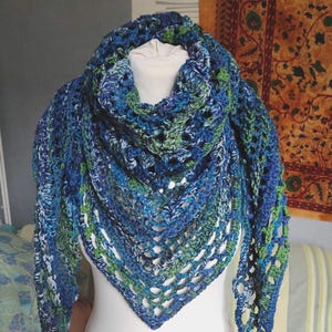 May include: A handmade, open-weave shawl in shades of blue and green is displayed on a white mannequin. The shawl is crocheted with a variety of blue hues, accented with green, creating a textured, eye-catching accessory. The triangular shape is visible.