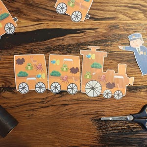 Könnte beinhalten: Ein orangefarbener Papiertrain mit Cartoon-Meeresbewohnern und einem Schaffner. Die Waggons zeigen Illustrationen von Fischen, Fröschen und Oktopussen. Eine schwarze Schere und eine Garnrolle liegen ebenfalls auf der Holzoberfläche.