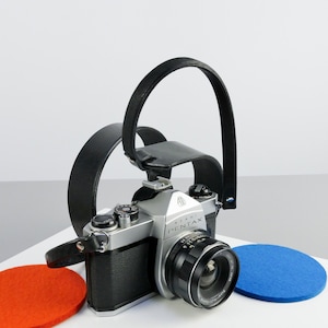 Puede incluir: Una cámara de película de 35 mm de color plateado y negro con una correa de cuero negra. La cámara tiene un objetivo con un anillo plateado y el texto "ASAHI PENTAX" impreso en la parte delantera.