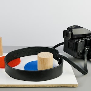 Puede incluir: Una correa de cámara de cuero negro con un cilindro de madera y círculos de colores sobre una superficie blanca.