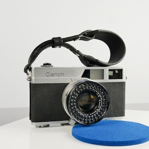 Puede incluir: Una cámara Canon plateada con una correa de cuero negra. La cámara tiene un objetivo negro con un anillo plateado alrededor. La cámara está sobre una superficie azul.