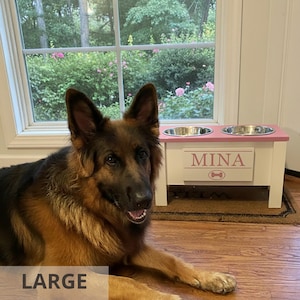 German shepherd beside large elevated dog feeder “MINA.” Custom raised wooden dog bowl stand with personalized engraved nameplate and removable stainless bowls. Farmhouse modern pet feeder supports healthy digestion and reduces strain on joints. Perfect for large breed dogs, handcrafted for durability, available in multiple colors and customizable designs. Stylish and functional pet furniture for every home.