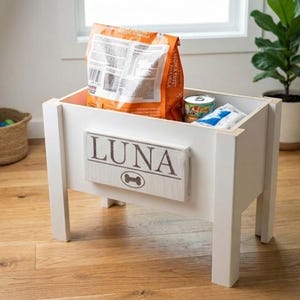 May include: White wooden pet food storage container with the name "LUNA" and a dog bone graphic. The container holds a bag of dog food, a can, and a packet. The container is elevated on four legs.