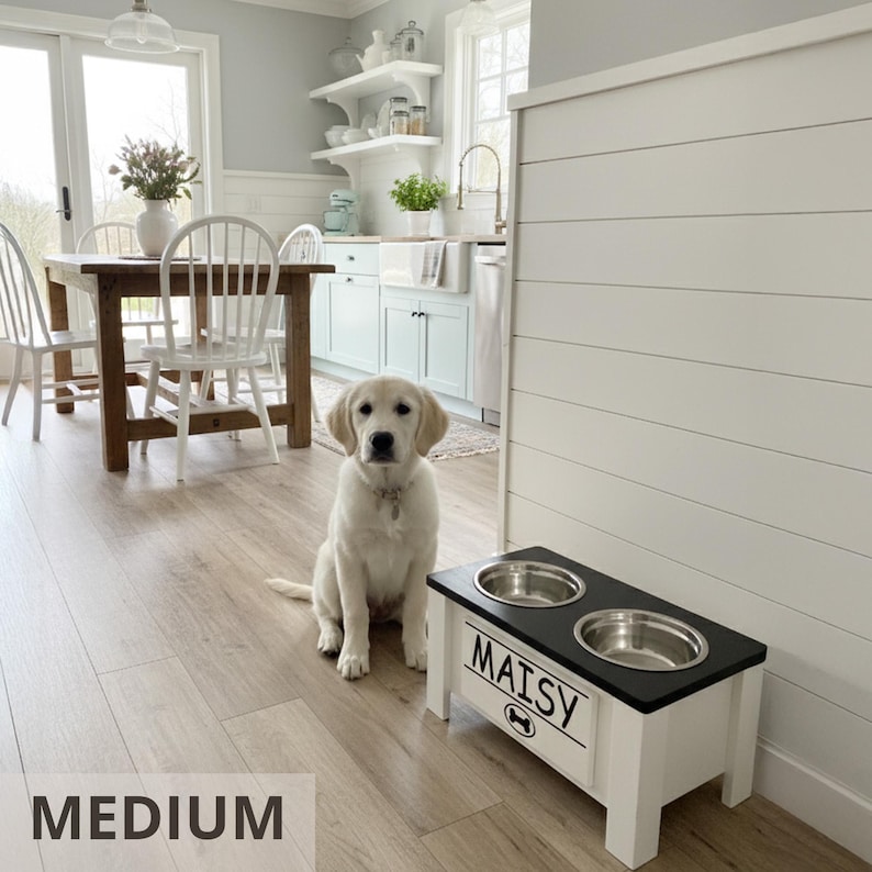 White dog sitting next to medium elevated feeder “MAISY.” Custom farmhouse wooden raised dog bowl stand with engraved nameplate, removable stainless bowls, and interchangeable lid options. Improves posture and digestion for medium size dogs. Personalizable design with stylish farmhouse look to match modern homes. Durable handcrafted feeder available in multiple sizes and colors for every breed.
