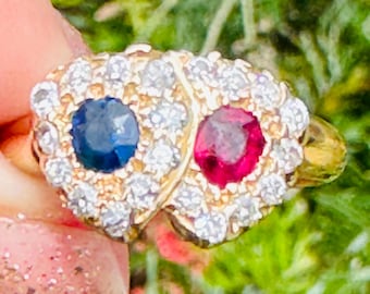 May include: A gold ring featuring two gemstone clusters. One cluster has a blue gemstone, the other a red gemstone, each surrounded by small, clear stones. The ring is held against a backdrop of green foliage.