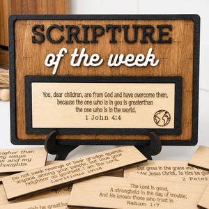 May include: A rectangular wooden display sign with the black and white text SCRIPTURE of the week and a central panel featuring 1 John 4:4. Below the sign are several smaller wooden rectangular cards, each engraved with different Bible verses.