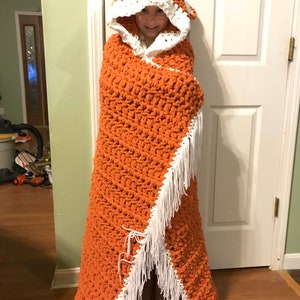 Puede incluir: Una manta de ganchillo con capucha, de color naranja con ribetes blancos. La manta tiene un borde de flecos y est&aacute; hecha con un punto texturizado.