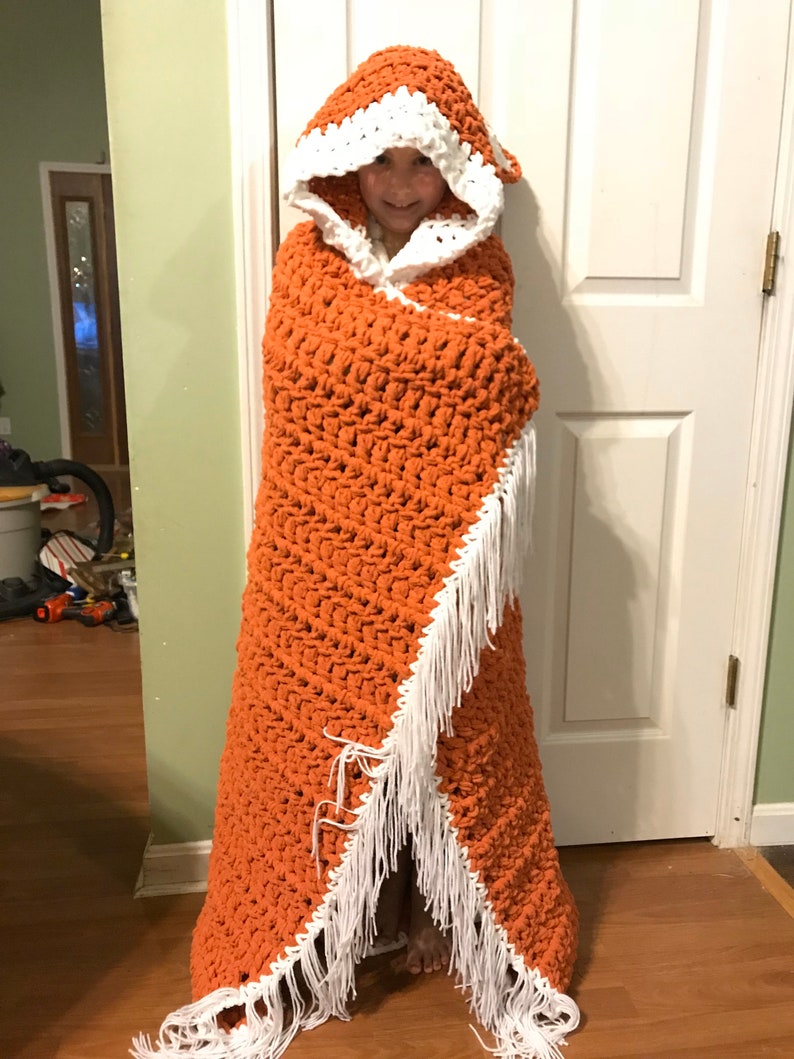 Puede incluir: Una manta de ganchillo con capucha, de color naranja con ribetes blancos. La manta tiene un borde de flecos y est&aacute; hecha con un punto texturizado.