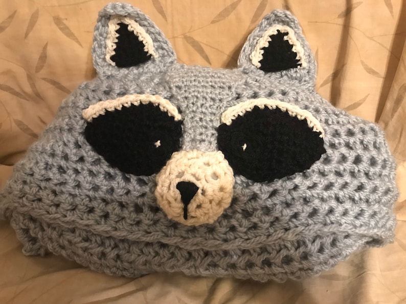 Op de afbeelding: Grijze gehaakte kussen in de vorm van een wasbeer met zwarte ogen, een witte neus en witte accenten op de oren.