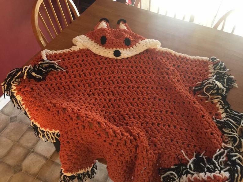 Puede incluir: Una manta de capucha de zorro de ganchillo hecha con hilo naranja, blanco y negro. La manta tiene un borde de flecos y una cara de zorro en la capucha.