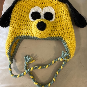 Puede incluir: Un gorro de ganchillo amarillo hecho a mano con forma de cabeza de perro. El gorro presenta orejas negras caídas, ojos blancos y negros y una nariz negra. El gorro tiene una corbata trenzada gris y amarilla.