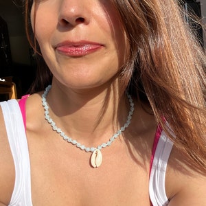 Op de afbeelding: Een lichtblauwe kralenketting met een witte schelp hanger.
