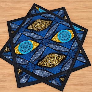 Puede incluir: Dos manteles individuales cuadrados de tela con un patrón geométrico repetitivo en tonos azul, negro y dorado. El diseño incluye formas estilizadas similares a ojos con iris azules y detalles dorados, bordeados por un ribete de tela negra.