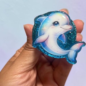 May include: A glossy, die-cut sticker or acrylic charm featuring a cartoon dolphin with large, expressive eyes. The dolphin is white with soft blue and pink gradients, surrounded by a thick, dark blue border filled with sparkling glitter.