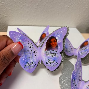 May include: Purple butterfly-shaped memorial keepsakes with a photo of a person named Tanya. The butterflies have glitter edges and include the dates 03/30/1976 and 01/24/2026, along with the words "Sunrise" and "Sunset". The text "Gone from sight, but never from our hearts" is also included.