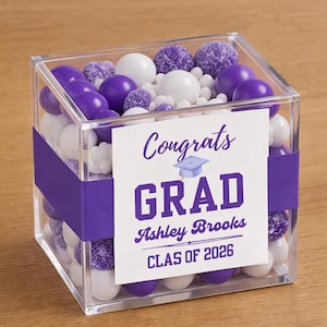 May include: A clear acrylic box filled with purple, white, and sugared candies. A purple ribbon wraps around the box, holding a white card that reads "Congrats GRAD Ashley Brooks CLAS OF 2026" in purple text. Graduation themed.