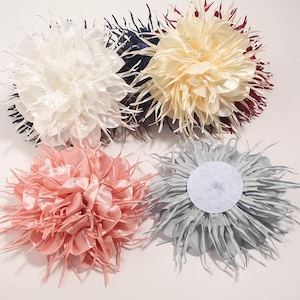 以下が含まれることがあります： 白、クリーム、ピンク、グレーの4色の布製フラワーヘアアクセサリー。各花は、繊細で羽のようなエッジと小さなキラキラした装飾が施された、中央のレイヤーデザインになっています。花は白い表面に配置されています。