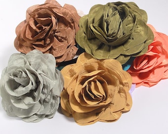 May include: A collection of large, fabric flower embellishments in various colors. The flowers include shades of brown, green, peach, gray, and gold. The petals are layered and textured, creating a realistic rose appearance. These could be used for crafts or fashion.