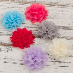 May include: Six fabric flowers in various colors, including red, pink, blue, gray, purple, and cream. The flowers are made of a delicate lace material and have a ruffled appearance. They are arranged on a white wooden surface.