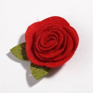 K&ouml;nnte beinhalten: Eine handgefertigte rote Filzrose mit zwei gr&uuml;nen Bl&auml;ttern. Die Rose steht in voller Bl&uuml;te, mit eng gewickelten Bl&uuml;tenbl&auml;ttern. Die Bl&auml;tter sind ein ged&auml;mpftes Gr&uuml;n und bilden einen Kontrast zur leuchtend roten Bl&uuml;te. Geeignet f&uuml;r Bastelarbeiten.